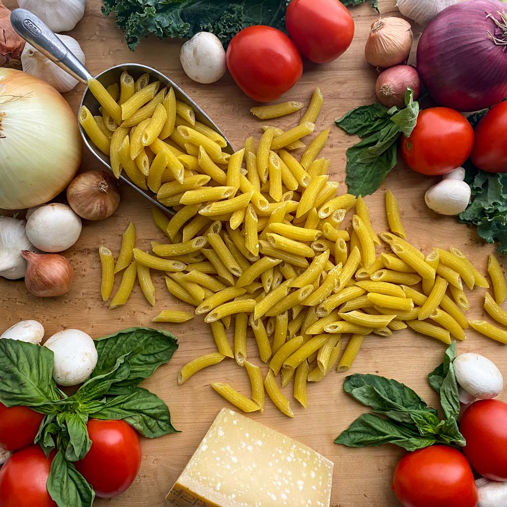 Fresh penne pasta on a cutting board surrounded by fresh tomatoes, basil whole onions, mushrooms, garlic, shallots and kale