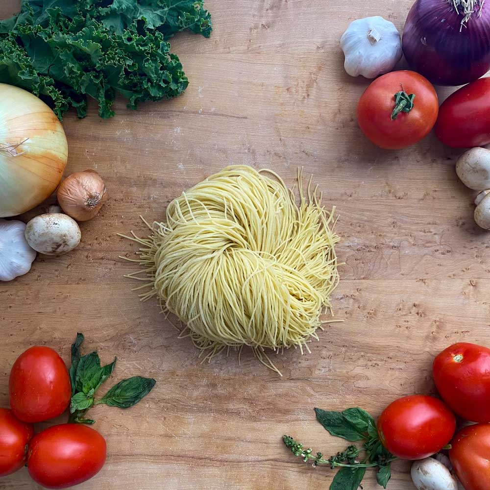 Fresh angel hair pasta on a cutting board surrounded by fresh tomatoes, basil whole onions, mushrooms, garlic, shallots and kale